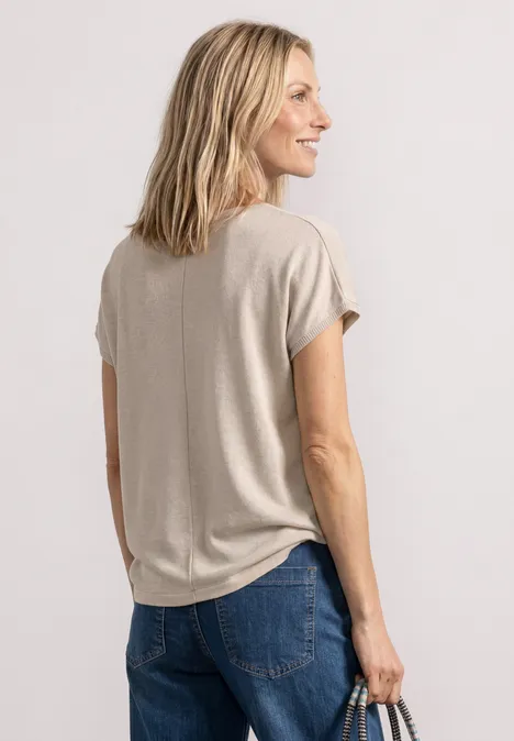Vrouw van achteren in beige T-shirt en spijkerbroek, met handtas in haar rechterhand.