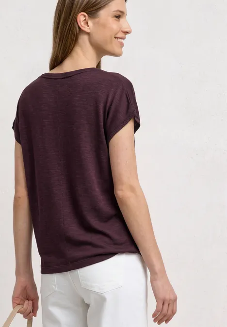 Dropped Shoulder Shirt mit Rundhals rustic rouge Frau trägt violettes T-Shirt und weiße Hose, von hinten fotografiert.