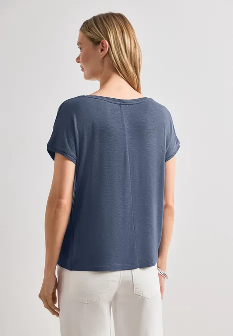 Dropped Shoulder Shirt mit Rundhals ashen blue Frau steht mit dem Rücken zur Kamera, trägt blaue Kurzarmbluse und weiße Hose.