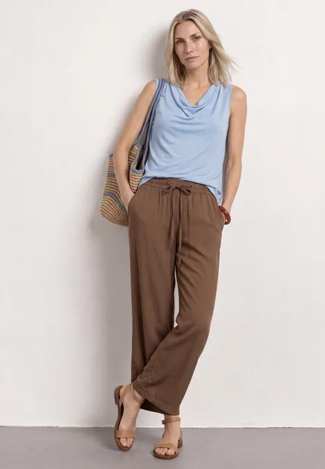 Silk-Look Top mit Wasserfall-Ausschnitt cashmere blue Frau lehnt in lässigem Outfit an weißer Wand: hellblaues Top, braune Hose, Sandalen und Strohtasche.