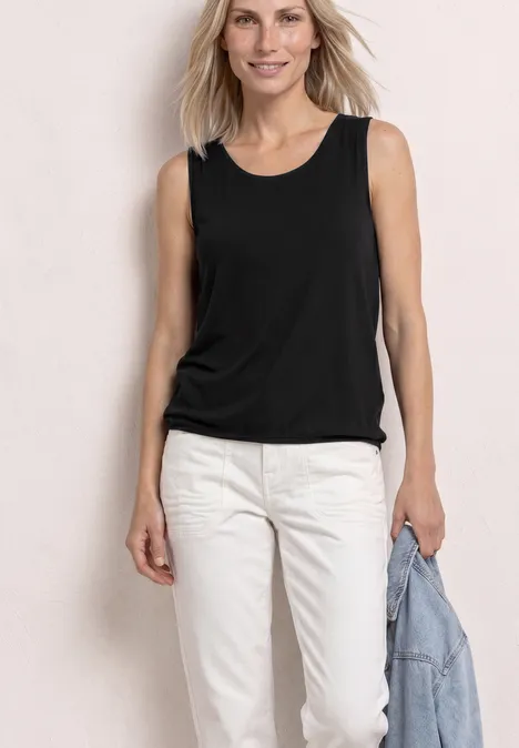 Frau in schwarzem ärmellosem Top und weißen Hosen, hält Jeansjacke in der Hand, vor hellem Hintergrund.