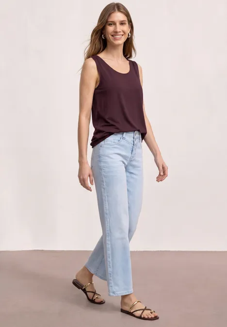 Frau in ärmellosem bordeauxfarbenen Top, weiten hellen Jeans und schlichten Riemchensandalen.