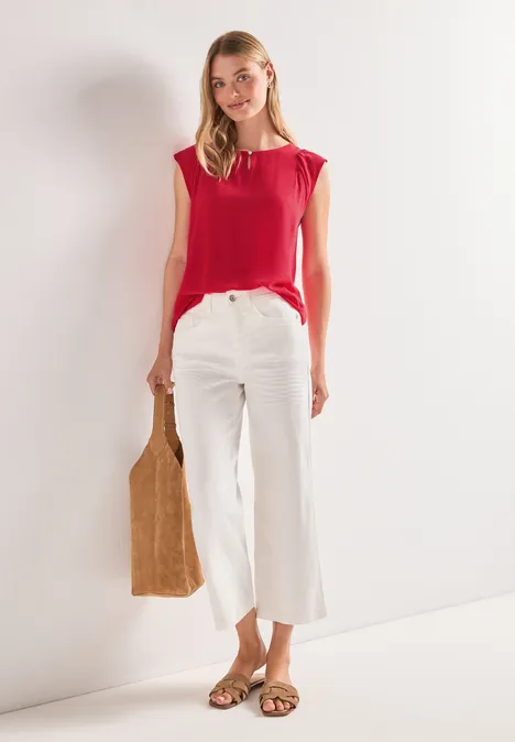 Vrouw in rood shirt en witte, wijdvallende broek, met bruine tas en platte sandalen.