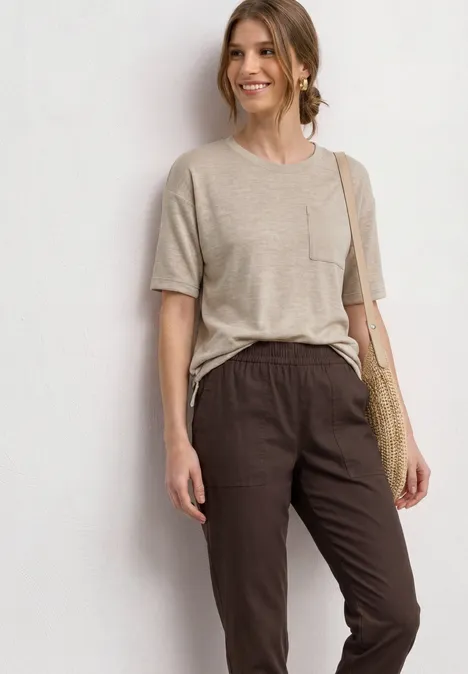Frau trägt beige T-Shirt, dunkle Stoffhose und Basttasche vor hellem Hintergrund.