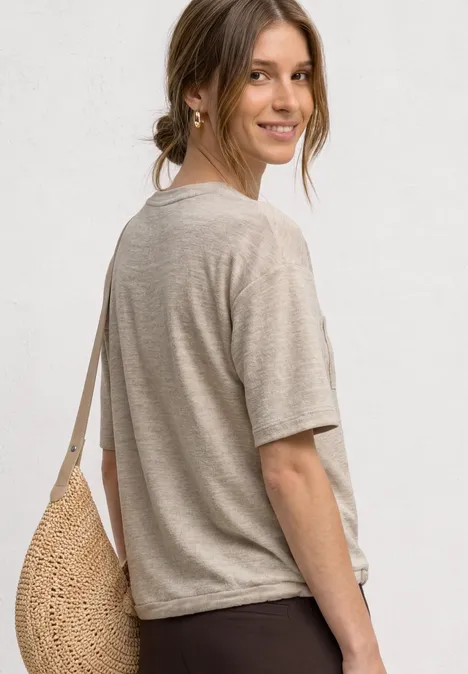 Junge Frau von der Seite mit beigem T-Shirt, Strohtasche über der Schulter vor weißem Hintergrund.