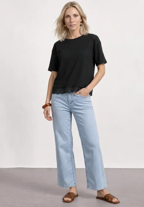 Frau in schwarzem T-Shirt, hellblauen Jeans und braunen Sandalen vor neutralem Hintergrund.