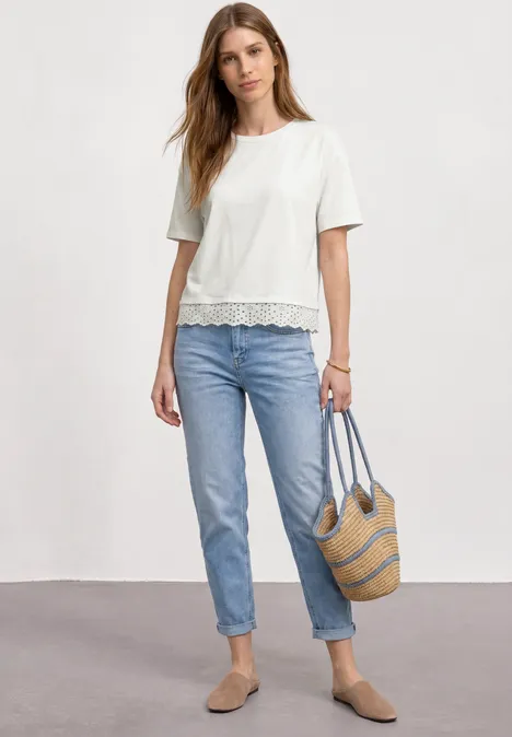 Frau trägt weiße Spitzenbluse, blaue Jeans, beigen Slipper und Strohtasche.