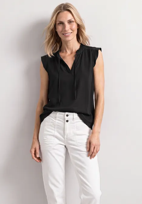 Vrouw voor witte muur met zwarte mouwloze blouse en witte broek.