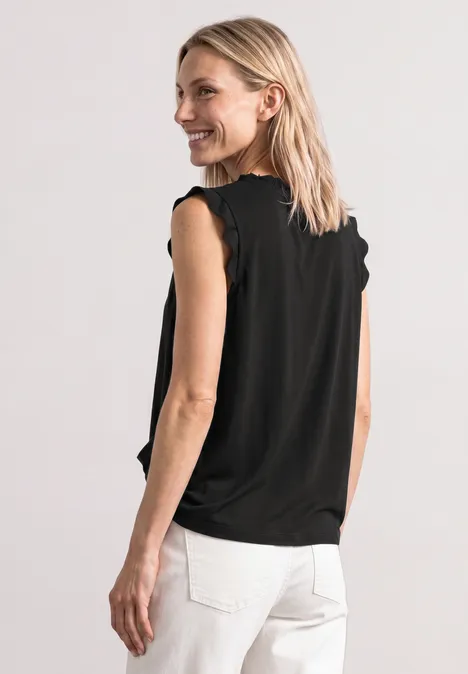 Vrouw van achter gefotografeerd in een zwarte mouwloze top en witte broek tegen lichte achtergrond.