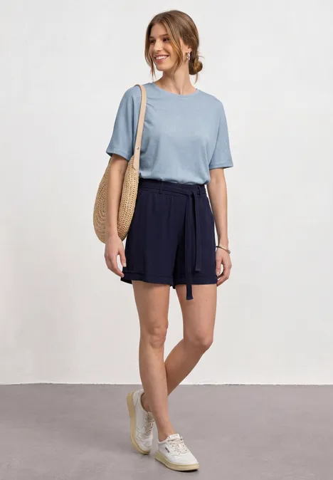 Frau in blauem T-Shirt, dunkelblauen Shorts, mit Strohtasche und weißen Sneakers.
