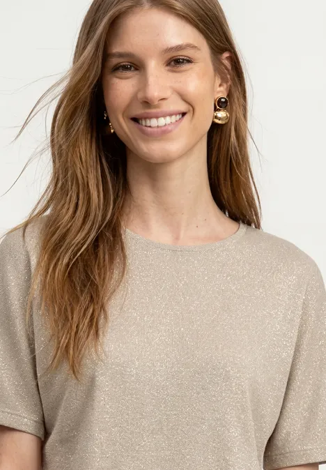 Frau trägt glitzernden beigen Pullover, lange braune Haare und goldene Ohrringe.