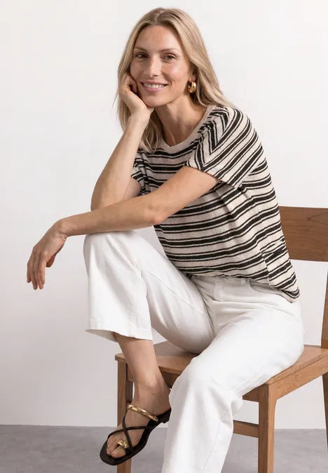 T-shirt en tricot avec col rond Black Frau sitzt auf schlichtem Holzstuhl, trägt gestreiftes Shirt, weiße Hosen und schwarze Sandalen.