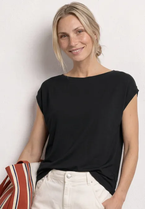 Dropped Shoulder Shirt mit Raffung Black Eine Frau trägt ein schwarzes, locker sitzendes T-Shirt mit hochgekrempelten Ärmeln und weiße Hosen.