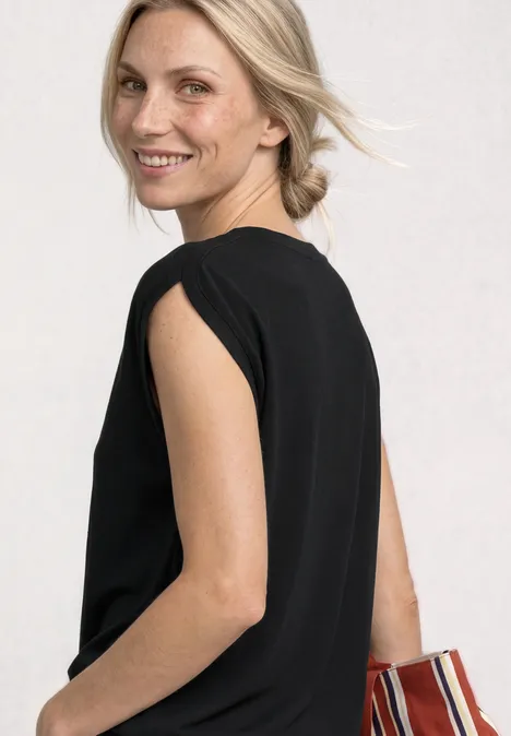 Dropped Shoulder Shirt mit Raffung Black Eine Frau in schwarzem, ärmellosem Oberteil, seitlich gedreht, mit gestreifter Tasche.