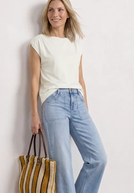 Frau mit weißem T-Shirt, hellblauer Jeans und gestreifter Tasche vor weißem Hintergrund.