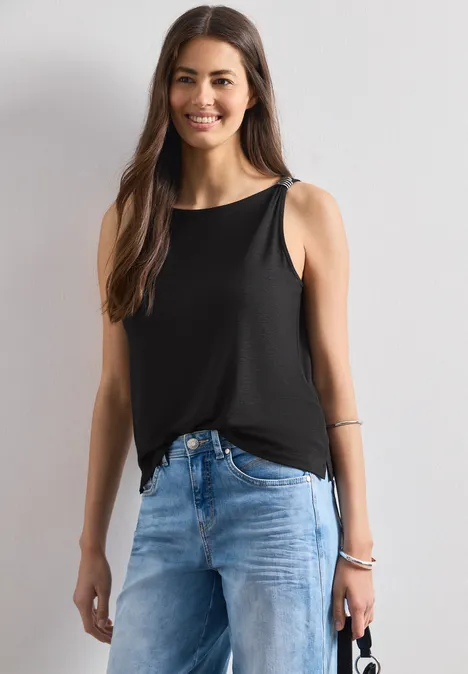 Eine Frau trägt ein schwarzes ärmelloses Top und blaue Jeans, hält eine kleine Tasche in der linken Hand.