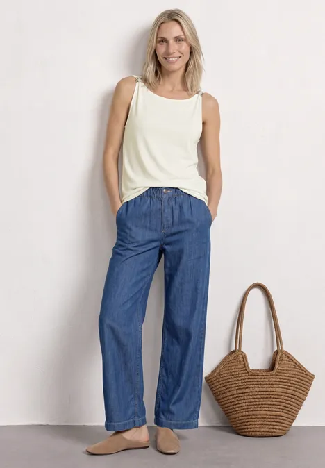 Frau in weißem Tanktop und weitem Jeans, neben Korbtasche stehend.