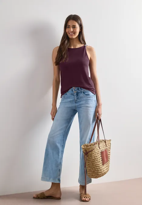 Frau vor neutralem Hintergrund in lässigem Look: dunkles Top, weite Jeans, Sandalen und Strohtasche.