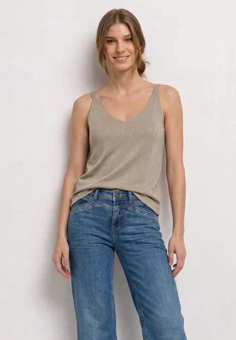 Top avec col en V et détails scintillants clay sand Femme en débardeur beige et jeans bleu devant un mur blanc.