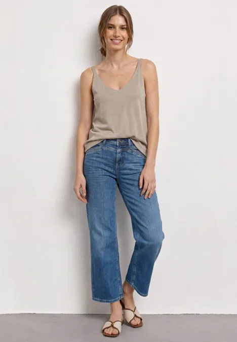 Top avec col en V et détails scintillants clay sand Femme en débardeur beige, jean court large bleu et sandales, posée devant un mur blanc.