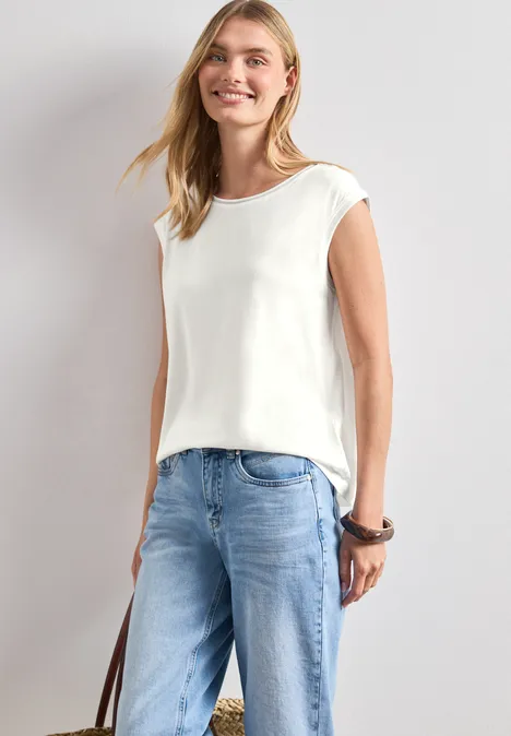 Schlanke Frau mit weißem ärmellosem Top und blauen Jeans, hält eine Tasche, vor hellem Hintergrund.