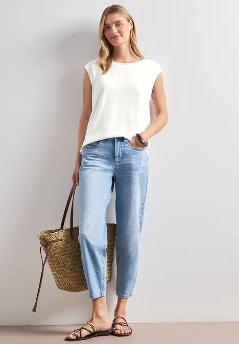 Frau in weißem Oberteil, hellen Jeans und schwarzen Sandalen trägt eine geflochtene Strohtasche.