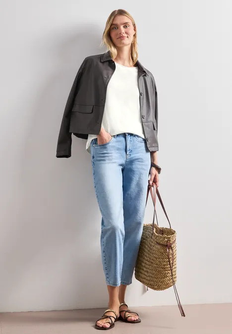 Frau mit weißem Top, dunkler Jacke über den Schultern, hellen Jeans, Sandalen und geflochtener Basttasche