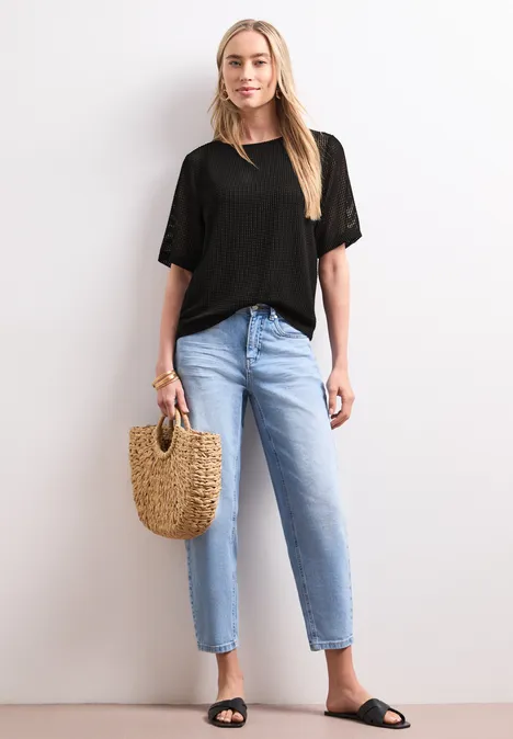 Eine schlanke Frau trägt ein schwarzes T-Shirt, helle Jeans, schwarze Sandalen und hält eine Basttasche.