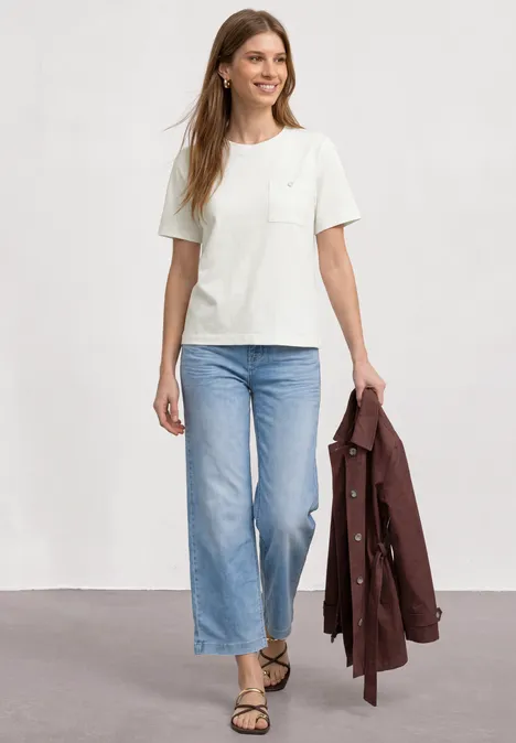 T-Shirt mit Rundhals und Brusttasche off white Person trägt weißes T-Shirt, hellblaue, weite Jeans, Sandalen und hält eine braune Jacke.