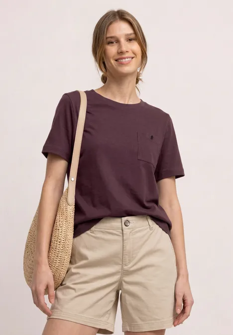 Eine stehende Person mit braunem T-Shirt, beigen Shorts und Strohtasche vor hellem Hintergrund.