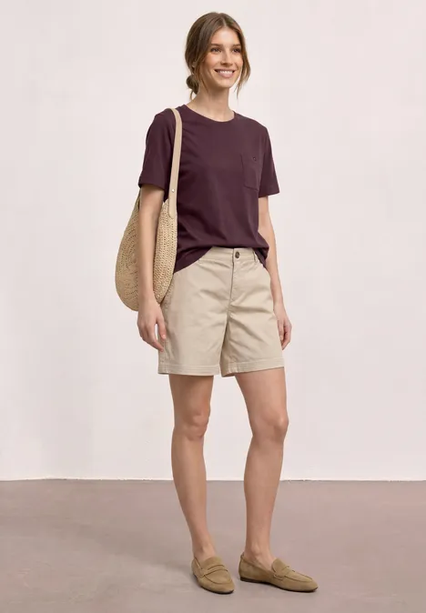 Person in legerer Kleidung: dunkelrotes T-Shirt, beige Shorts, Loafers und Korbtasche.