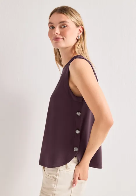 Seitlich fotografierte Frau mit ärmellosem lila Top und heller Hose vor hellem Hintergrund.