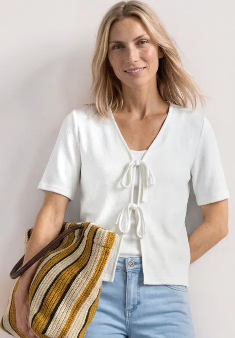 Veste en maille avec rubans off white Femme en haut blanc à manches courtes avec nœuds, jean bleu clair et sac rayé en tissu.