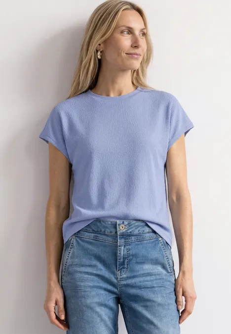 Frau trägt hellblaues T-Shirt und Jeans, steht vor weißem Hintergrund.