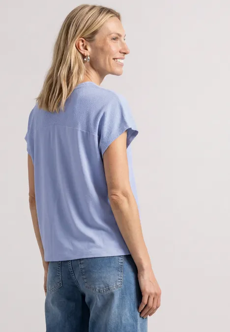 Eine Frau mit blonden Haaren trägt ein hellblaues T-Shirt und Jeans, im Profil, vor neutralem Hintergrund.