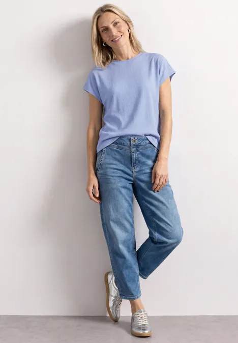 Frau in lässigem Outfit mit hellblauem T-Shirt, lockeren Jeans und Turnschuhen lehnt entspannt an Wand.