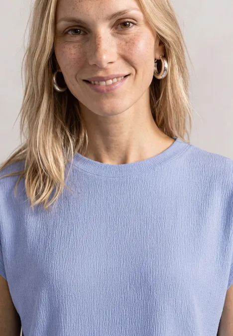 Eine Frau mit blonden Haaren trägt ein hellblaues ärmelloses Oberteil und silberne Creolen.