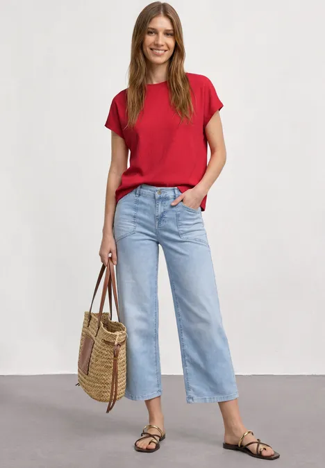 Frau trägt rotes T-Shirt, hellblaue, weite Cropped-Jeans, schwarze Sandalen und Bast-Shopper vor neutralem Hintergrund.