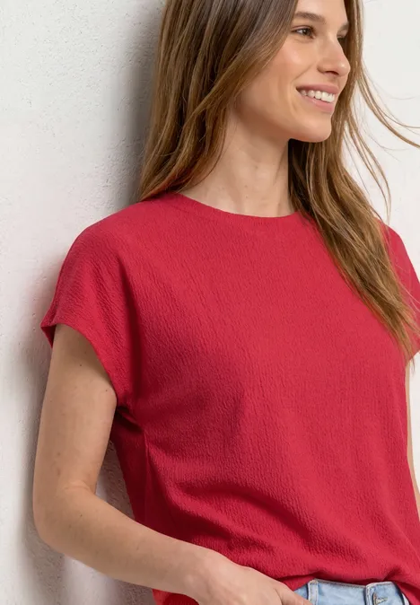 Frau lehnt an weißer Wand und trägt rotes Kurzarmshirt mit Jeans.