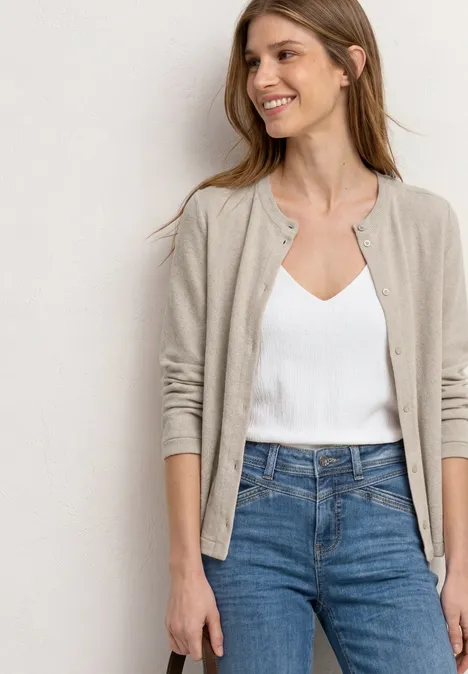 Vrouw in een wit topje, beige vest en blauwe jeans voor een neutrale achtergrond.