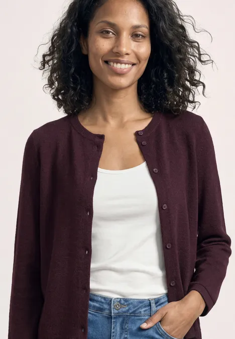Vrouw in bordeauxrode cardigan, witte top en spijkerbroek, krullend haar, hand in zak.
