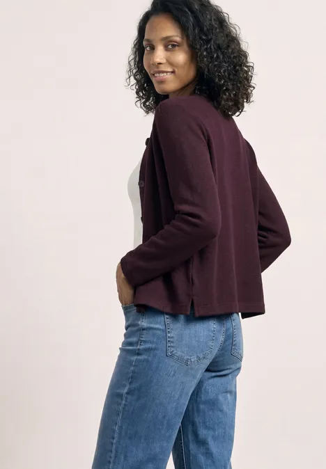 Vrouw met krullend haar draagt een bordeauxvest en blauwe jeans, zijaanzicht.