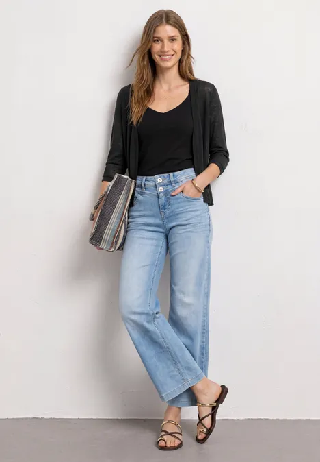 Frau in schwarzer Strickjacke, schwarzem Top, hellblauen Jeans und Sandalen, hält eine gestreifte Clutch.