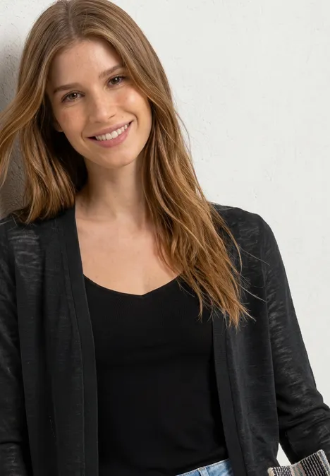 Person mit langen braunen Haaren, trägt schwarzen Pullover und schwarzen Cardigan vor weißem Hintergrund.