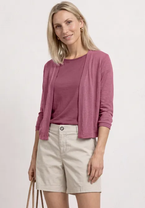 Femme blonde en t-shirt et cardigan roses avec short beige et sac assorti sur fond blanc.