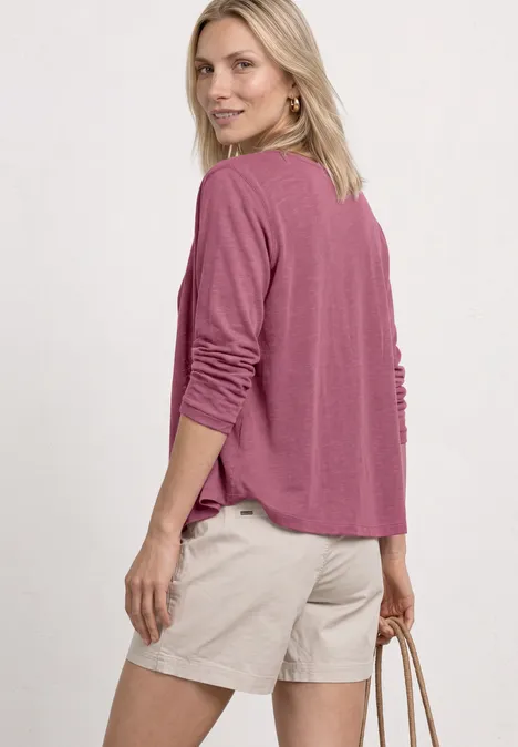 Femme de dos en haut rose à manches longues et short beige, tenant un sac.