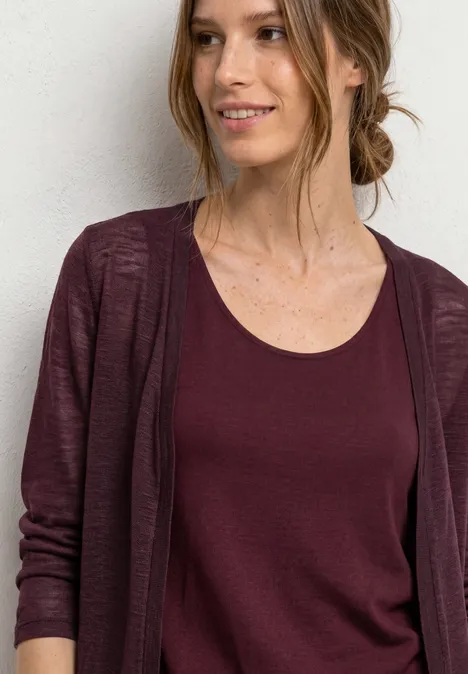 Eine lächelnde Frau mit lockerem, dunkelrotem Cardigan und passendem Top vor weißer Wand.