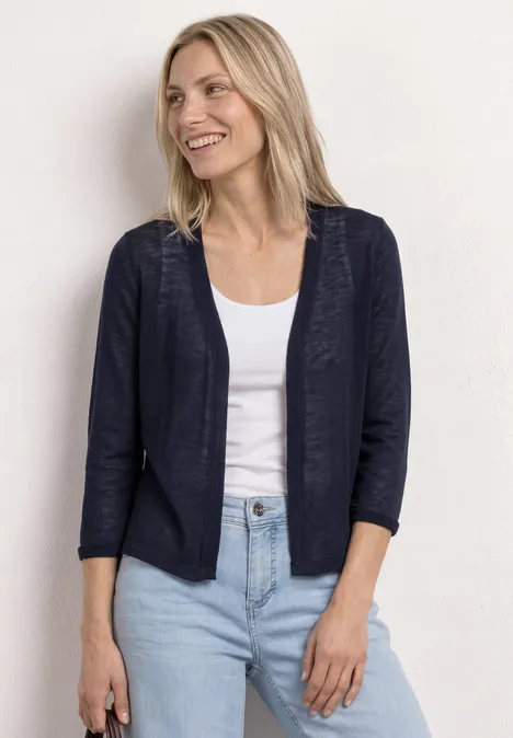 Frau in marineblauem Blazer über weißem Top und hellblauen Jeans vor hellem Hintergrund.