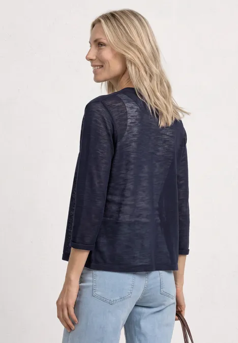 Eine Frau von hinten in dunkelblauer, leichter Strickjacke mit 3/4-Ärmeln und hellblauen Jeans, Tasche in der Hand.