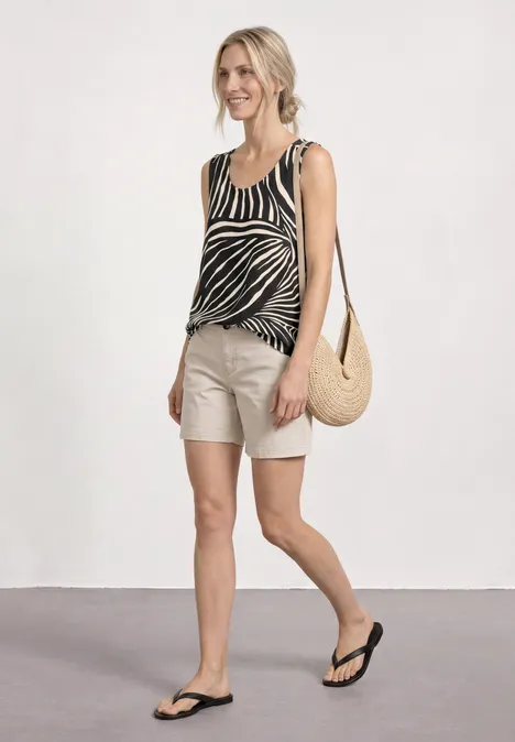 Frau trägt Zebra-Top, beige Shorts, Strohtasche und Flip-Flops, geht vor weißer Wand.